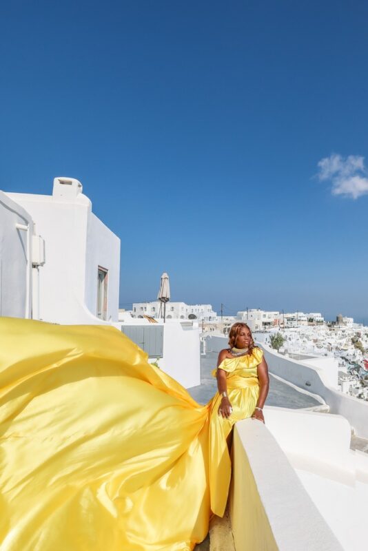 Black woman posing in a yellow flying dress in Imerovigli, Santorini. Posing ideas for a professional flying dress photoshoot.