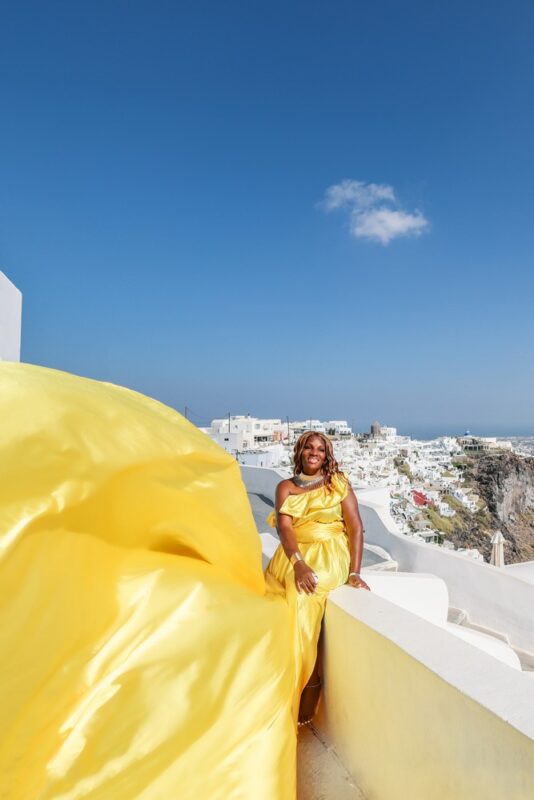 Black woman posing in a yellow flying dress in Imerovigli, Santorini. Posing ideas for a professional flying dress photoshoot.