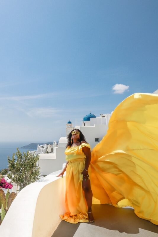 Black woman posing in a yellow flying dress in front of the blue dome church in Imerovigli, Santorini. Posing ideas for a professional flying dress photoshoot.